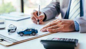 Driver reviews an auto insurance card beside a parked vehicle and vehicle registration documents.